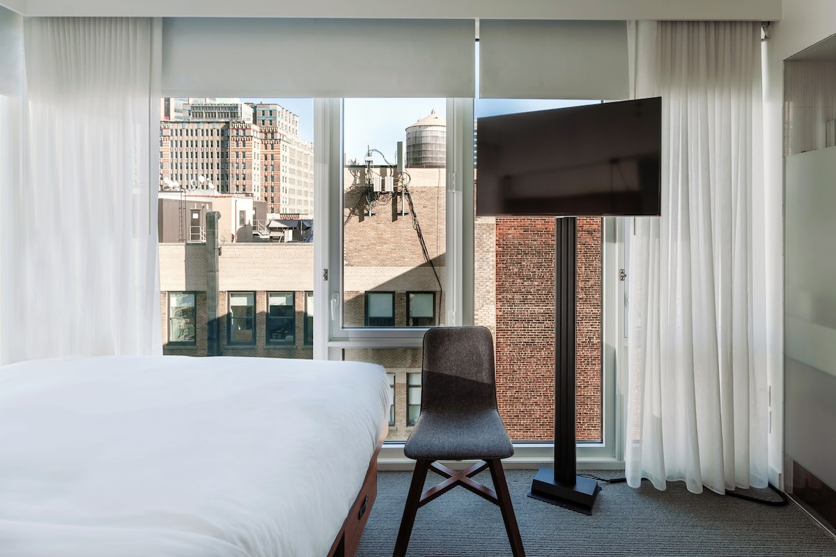 Ett hotellrum med en stor säng, en stol, en platt-TV och utsikt över stadsbilden genom ett stort fönster.