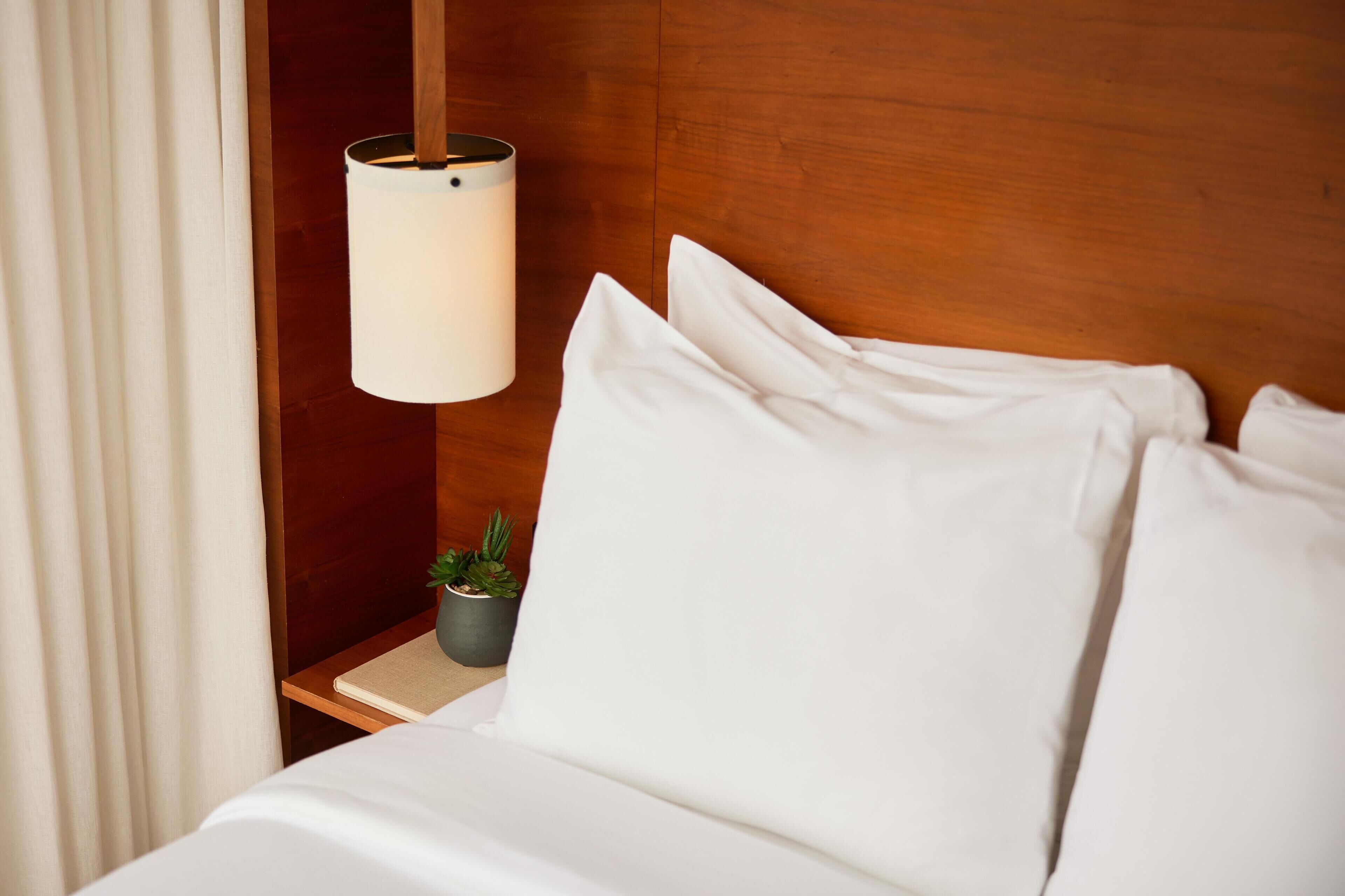 A neatly made bed with white linens, a bedside lamp, a small potted plant, and a wooden headboard.