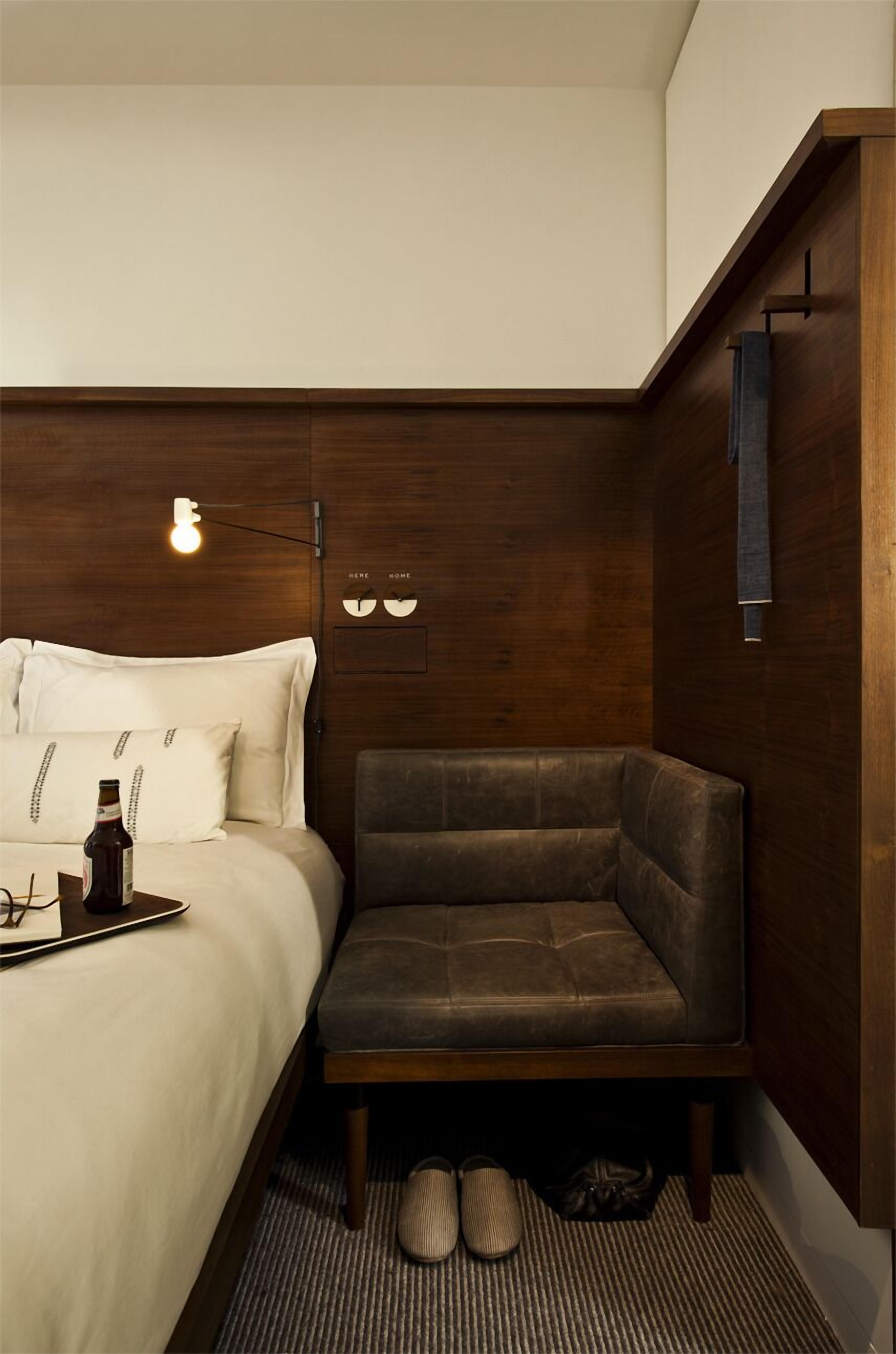 A compact hotel room with a bed, a small seating area, and a wall-mounted light fixture.