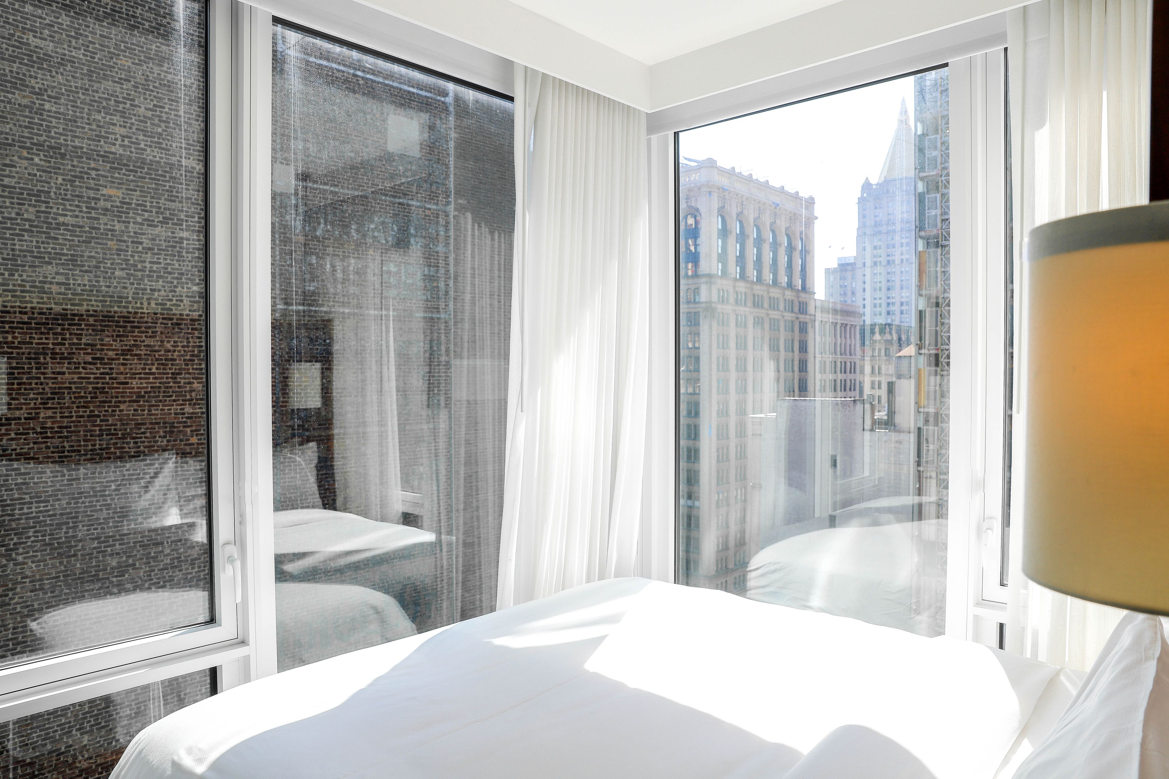 A hotel room with a large window, white curtains, and a view of city buildings.