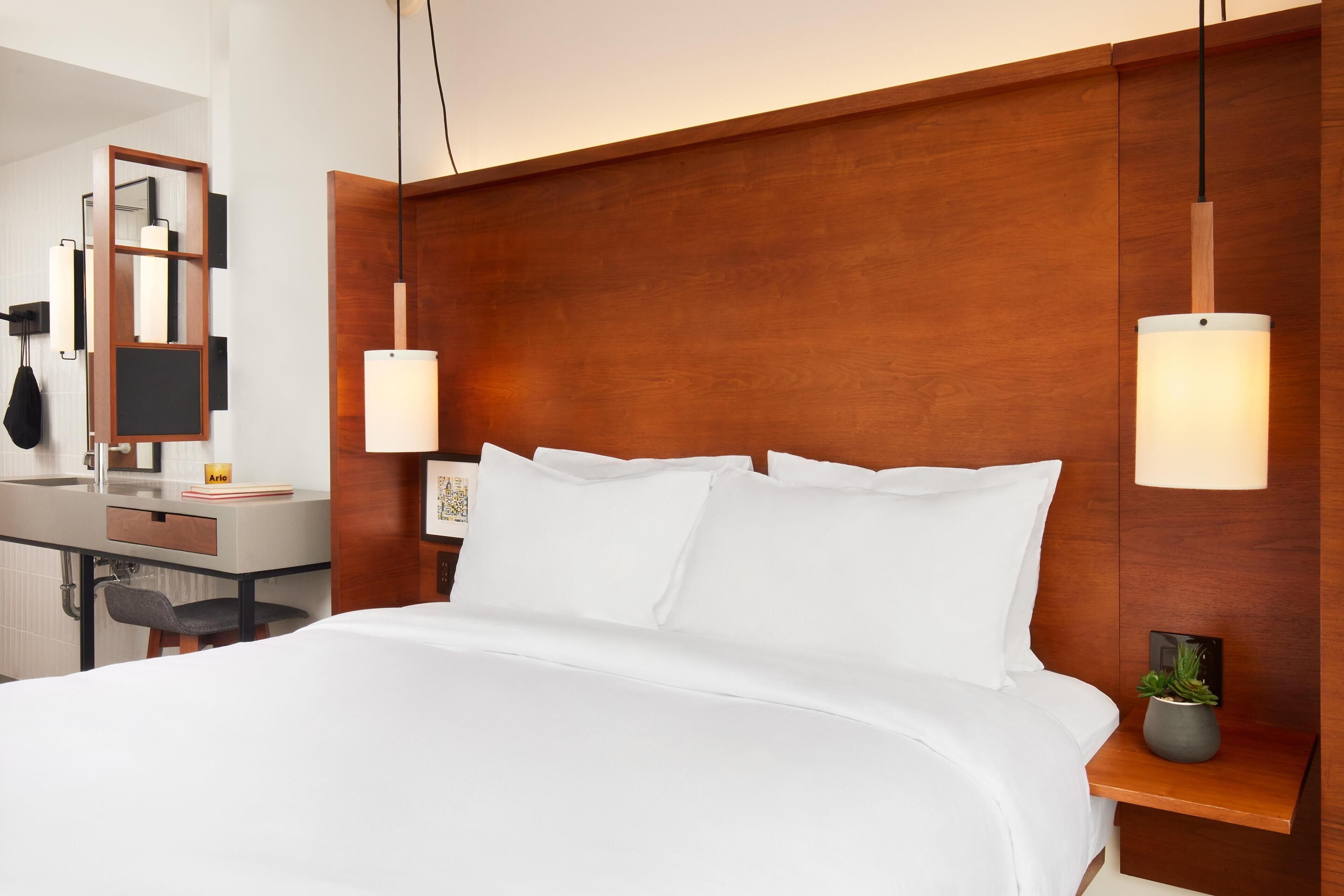 Una habitación de hotel con una cama grande, mesitas de noche, un escritorio con silla, un espejo y dos lámparas de pared.