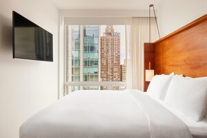 A hotel room with a large bed, a flat-screen TV mounted on the wall, and a view of a tall building through the window.