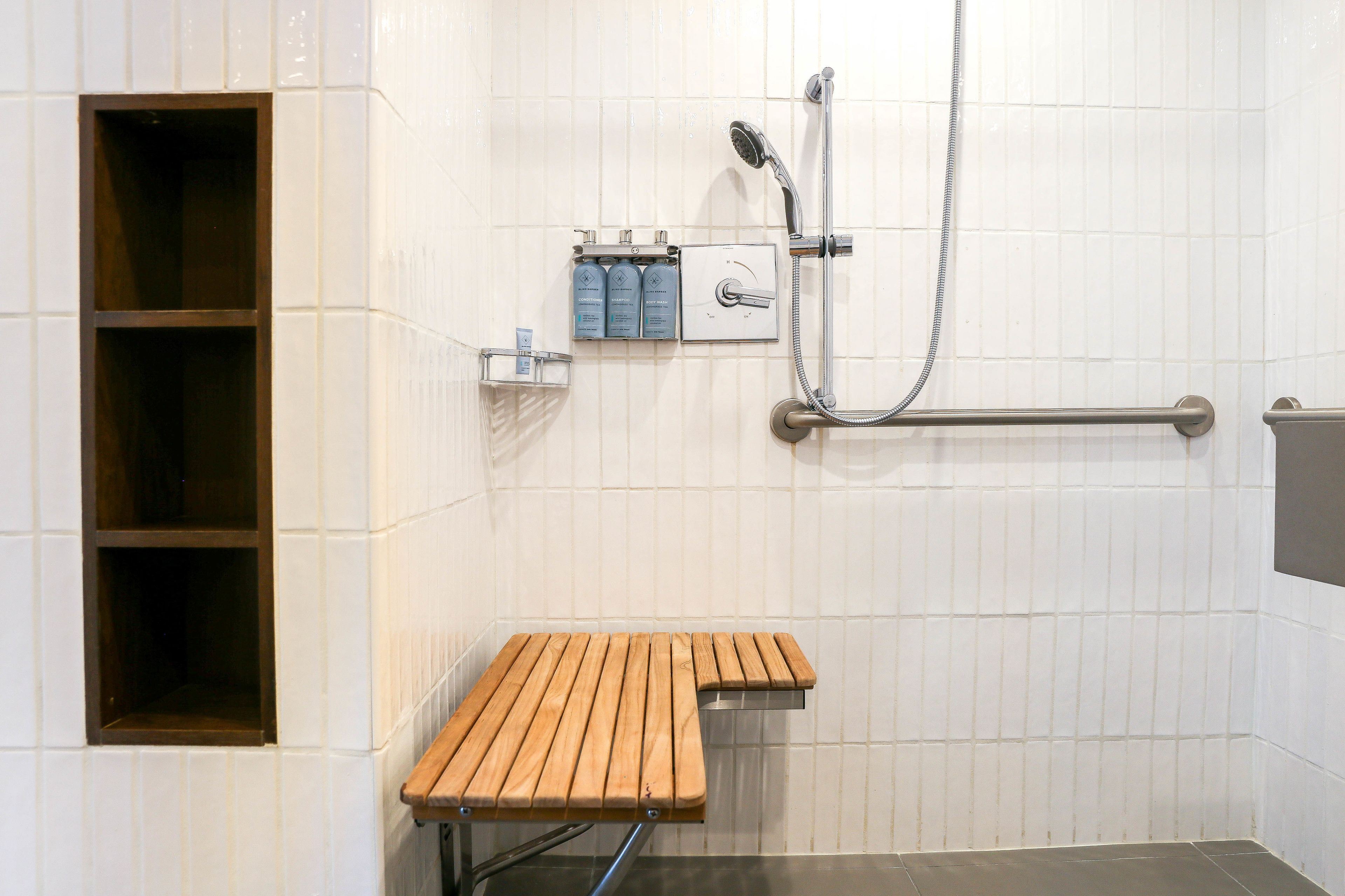 A bathroom with a wooden bench, a shower with a handheld showerhead, and a shelf with toiletries.