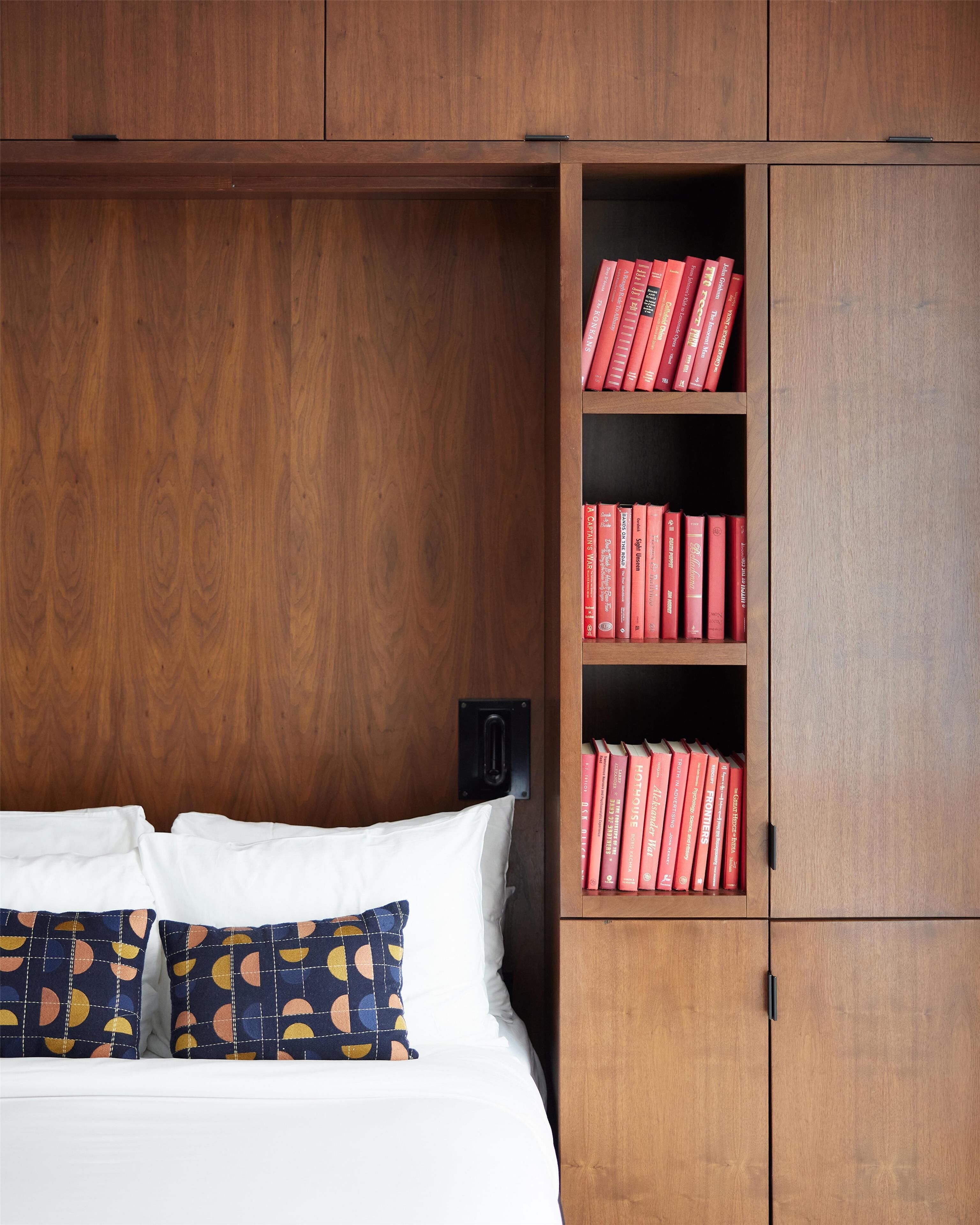 A bedroom with a wooden wardrobe, a bed with white bedding, and a shelf with red books.