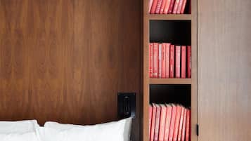 A bedroom with a wooden wardrobe, a bed with white bedding, and a shelf with red books.