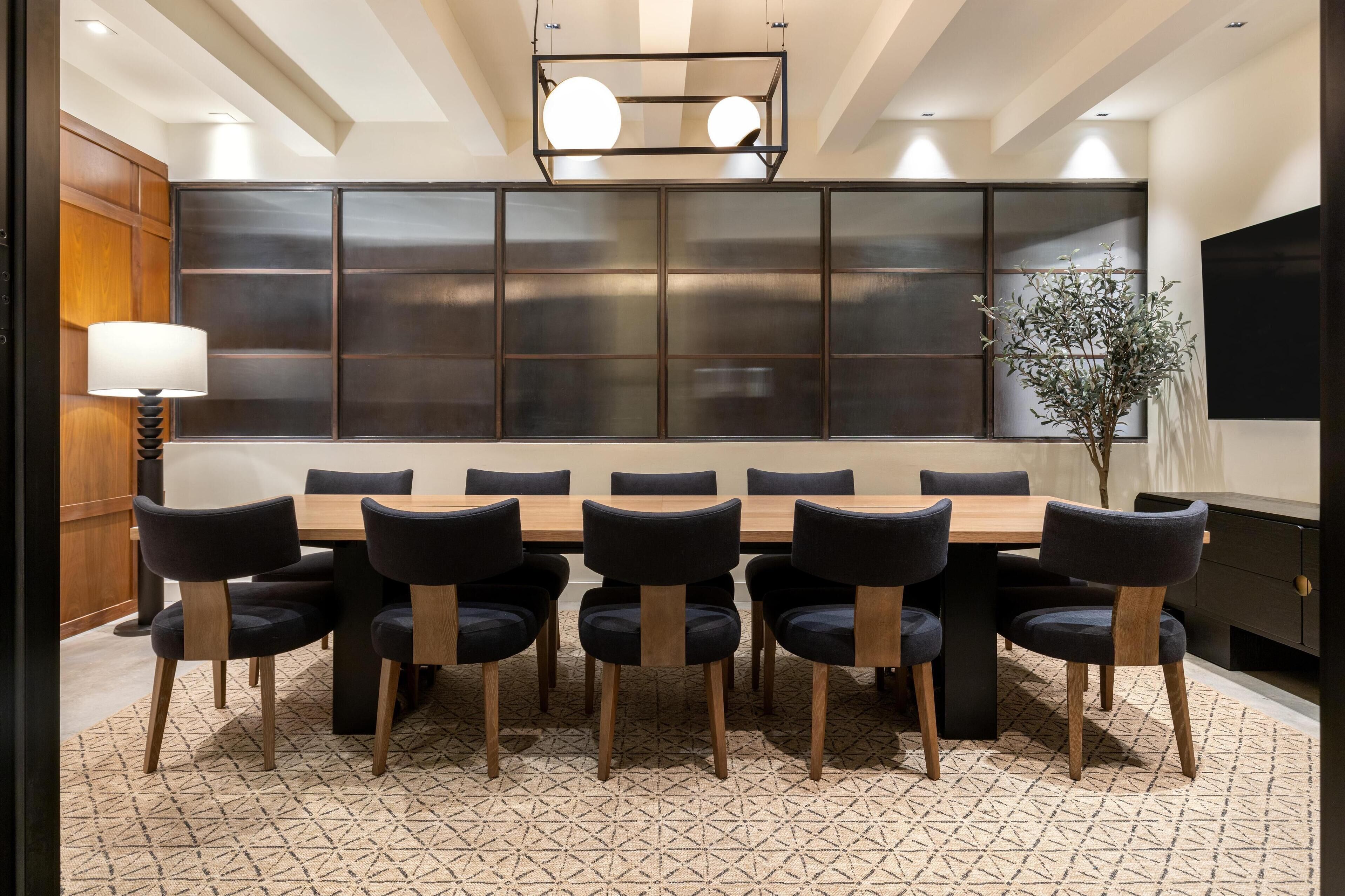 A dining area with a long wooden table surrounded by chairs, a large wall with a TV, and a floor lamp.