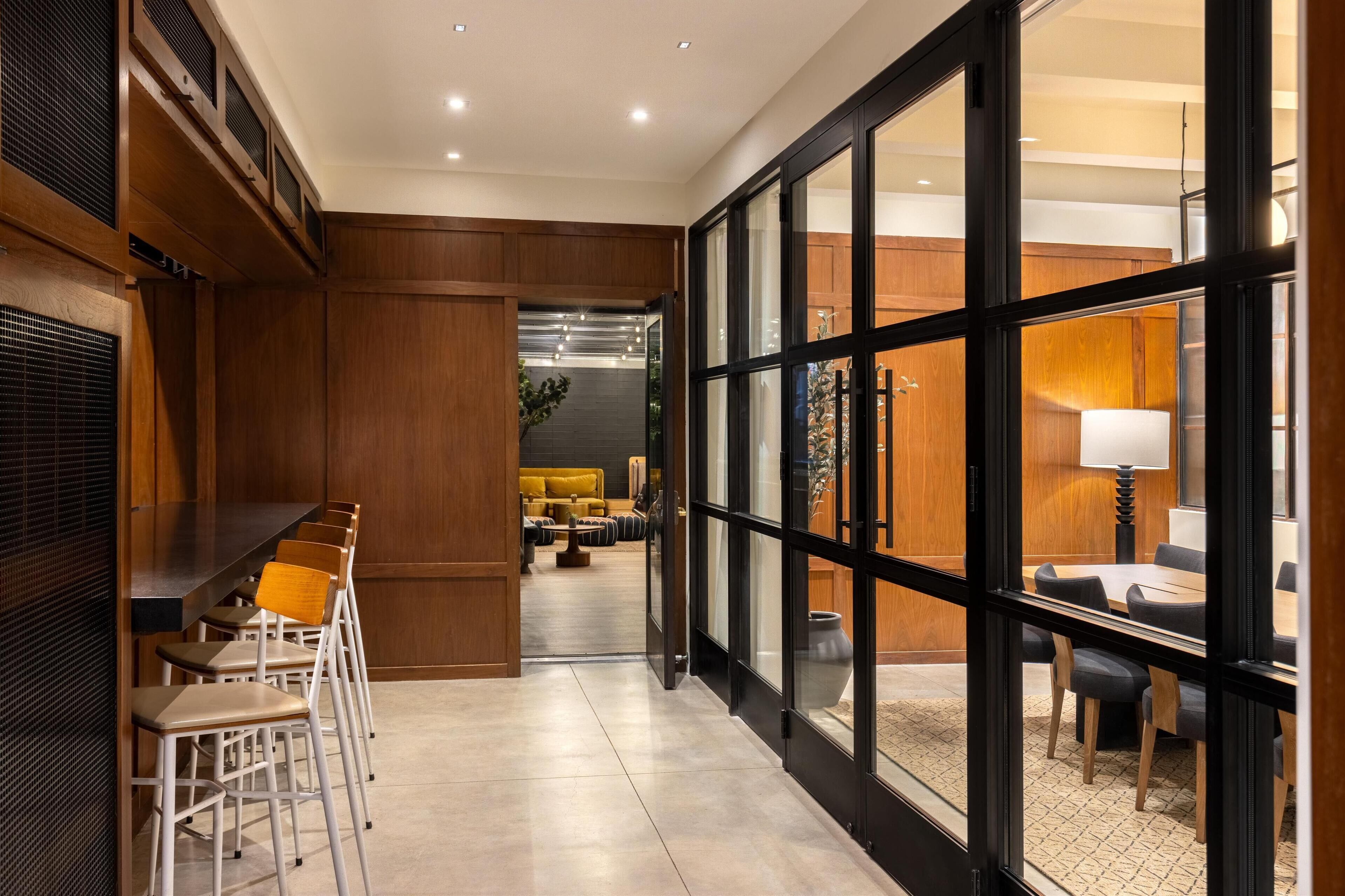 Un moderno vestíbulo de hotel con un largo pasillo, paneles de madera, una zona de estar con un sofá amarillo y un amplio ventanal con marcos negros.