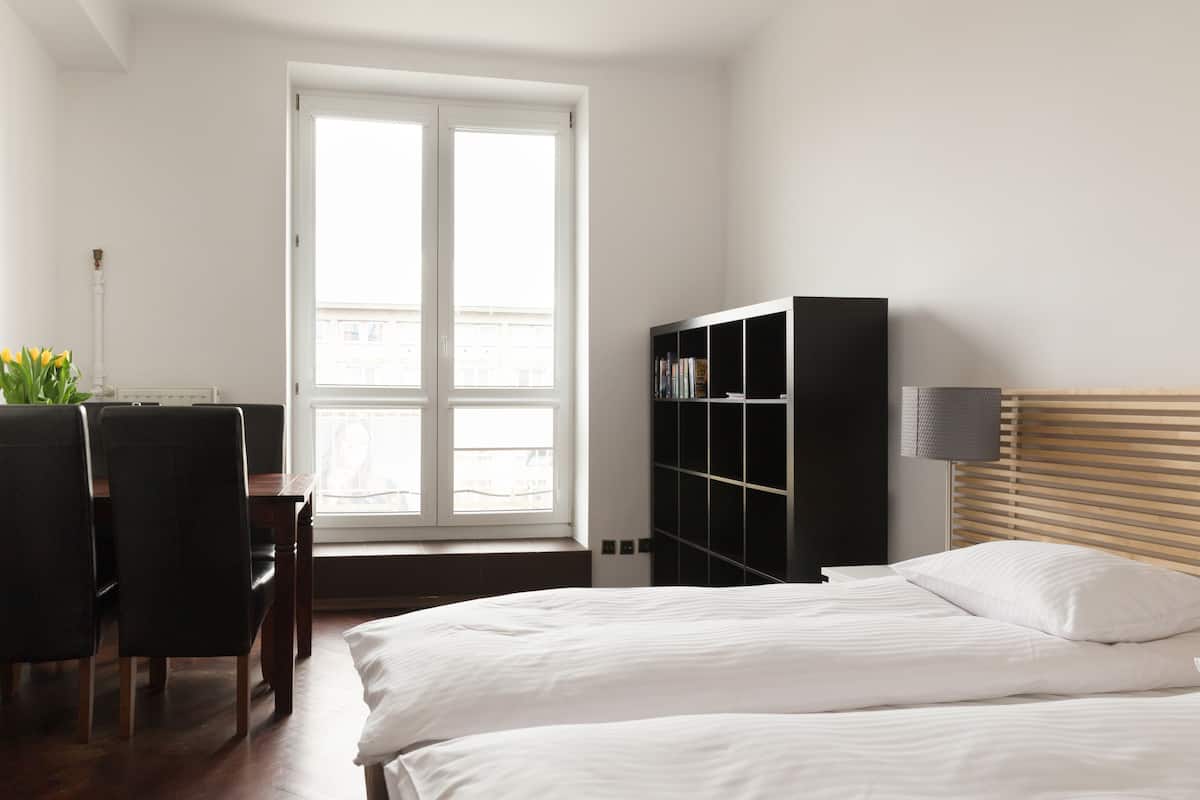 A hotel room with a large bed, a desk with a chair, a bookshelf, and a window with blinds.