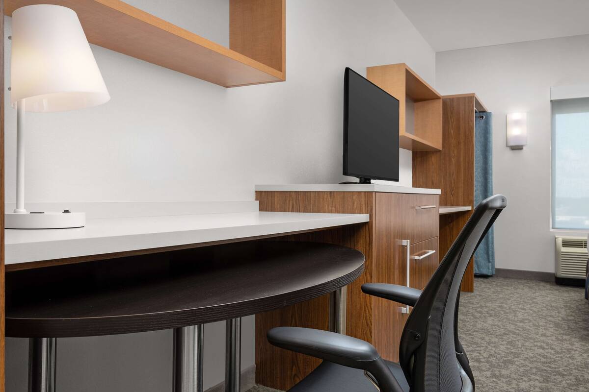 A modern hotel room with a desk, chair, TV, and built-in shelving.