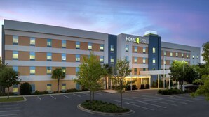 Un hotel moderno con un letrero que dice "HOME 2 SUITES by Hilton" y un estacionamiento con árboles.