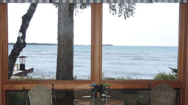 A room with a large window overlooking a body of water, two wicker chairs, a small round table with a wine glass, and a bird feeder outside. - Square Rigger Harbor Motel (Baileys Harbor)