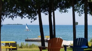 A lakeside view with two Adirondack chairs, a yellow table, and a dock with a sailboat in the distance. - Square Rigger Harbor Motel (Baileys Harbor)