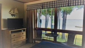 A hotel room with a bed, a TV on a wooden dresser, a balcony with a table and chairs, and a view of a lake. - Square Rigger Harbor Motel (Baileys Harbor)