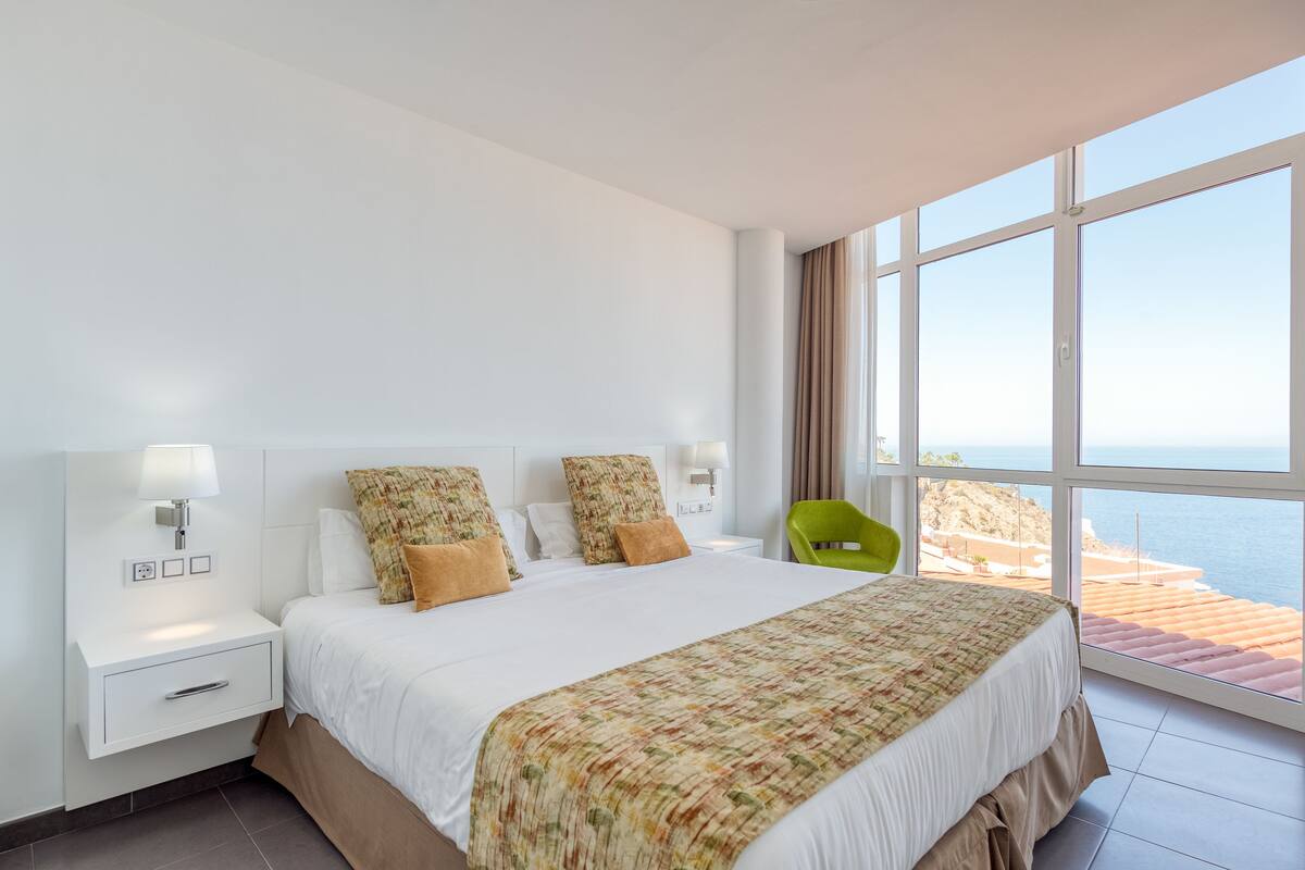 Een hotelkamer met een groot bed, een nachtkastje, een groene stoel en uitzicht op zee.