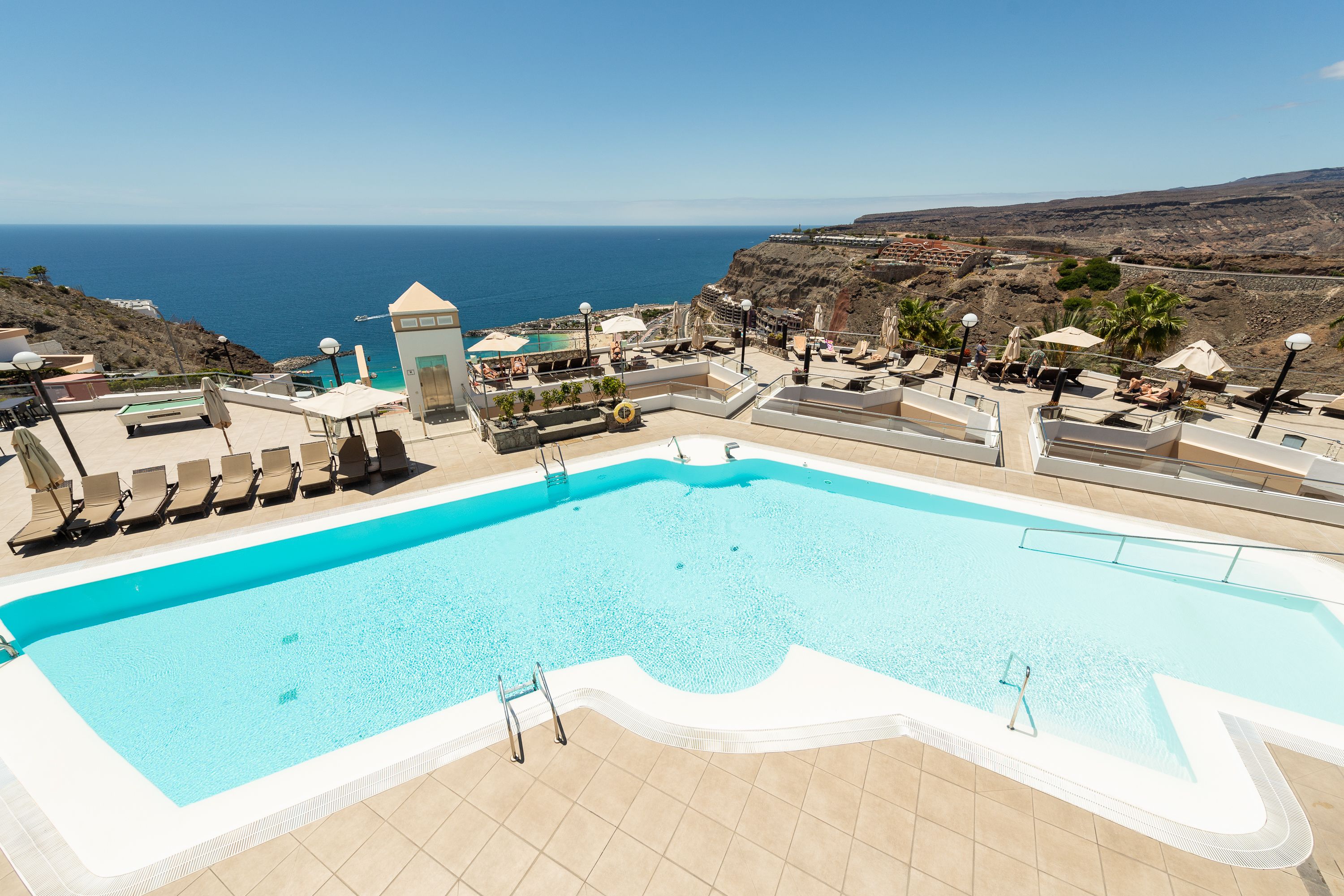 A swimming pool with a view of the ocean, sun loungers, and a small building.