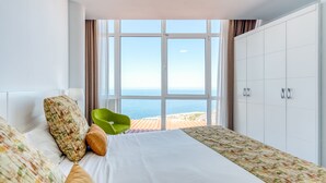 Une chambre d’hôtel avec une grande fenêtre, un lit avec une literie à motifs, un fauteuil vert et une armoire.