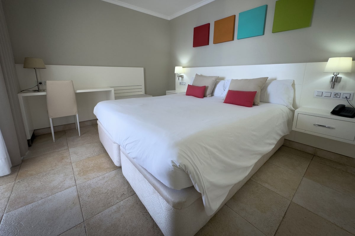 Hotelkamer met een groot bed, een bureau en een stoel. Drie lichtschakelaars op de muur en een telefoon op het nachtkastje.
