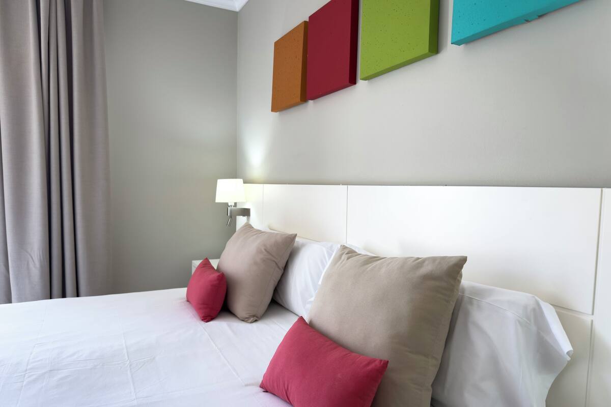 Een hotelkamer met een bed, wit beddengoed en een hoofdeinde. Daarboven hangt kleurrijke wanddecoratie.