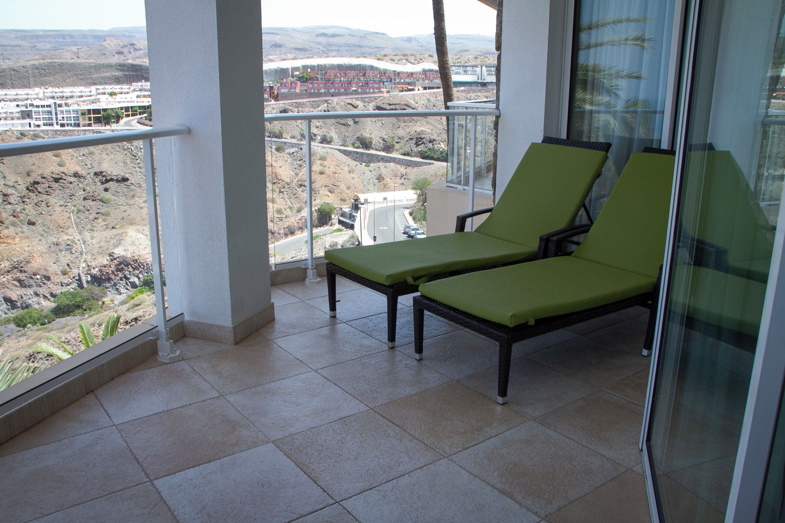 A balcony with two green lounge chairs, a glass railing, and a view of a construction site and hills in the distance.