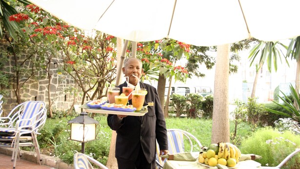 An outdoor dining area with chairs, a table with fruits, and a person serving food. - Sunshine Hotel (Asmara)