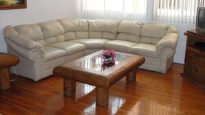 Un sofá seccional color beige, una mesa de centro de madera con un florero y un televisor sobre un mueble de madera.