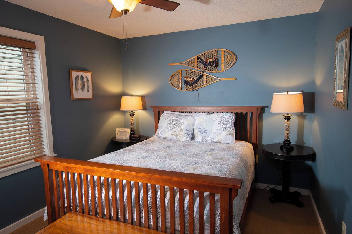 A bedroom with a wooden bed, two bedside tables with lamps, a ceiling fan, and a wall-mounted decorative item.
