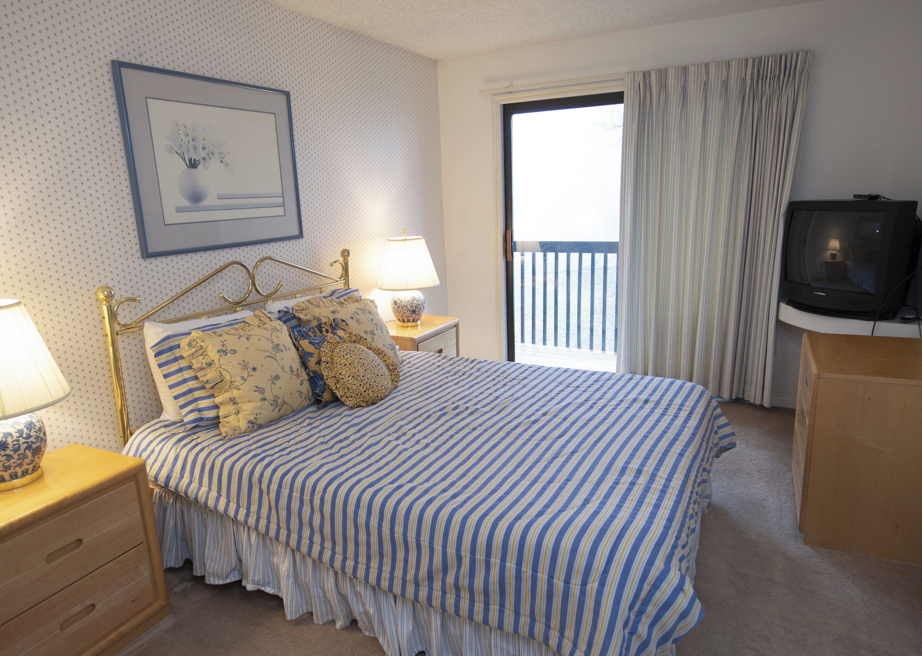 Un dormitorio con cama, mesitas de noche, lámpara, televisor y ventana con cortinas.