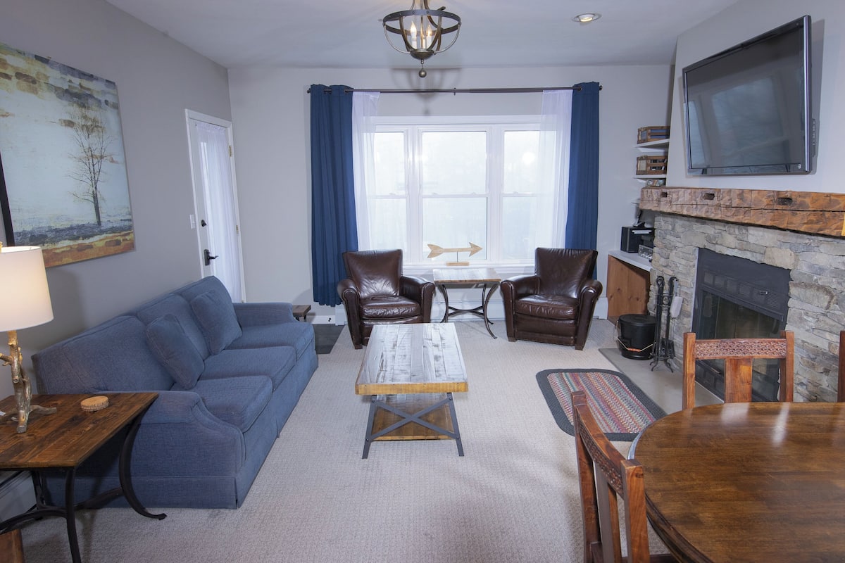 A living room with a blue sofa, a wooden coffee table, leather chairs, a dining table, and a stone fireplace.