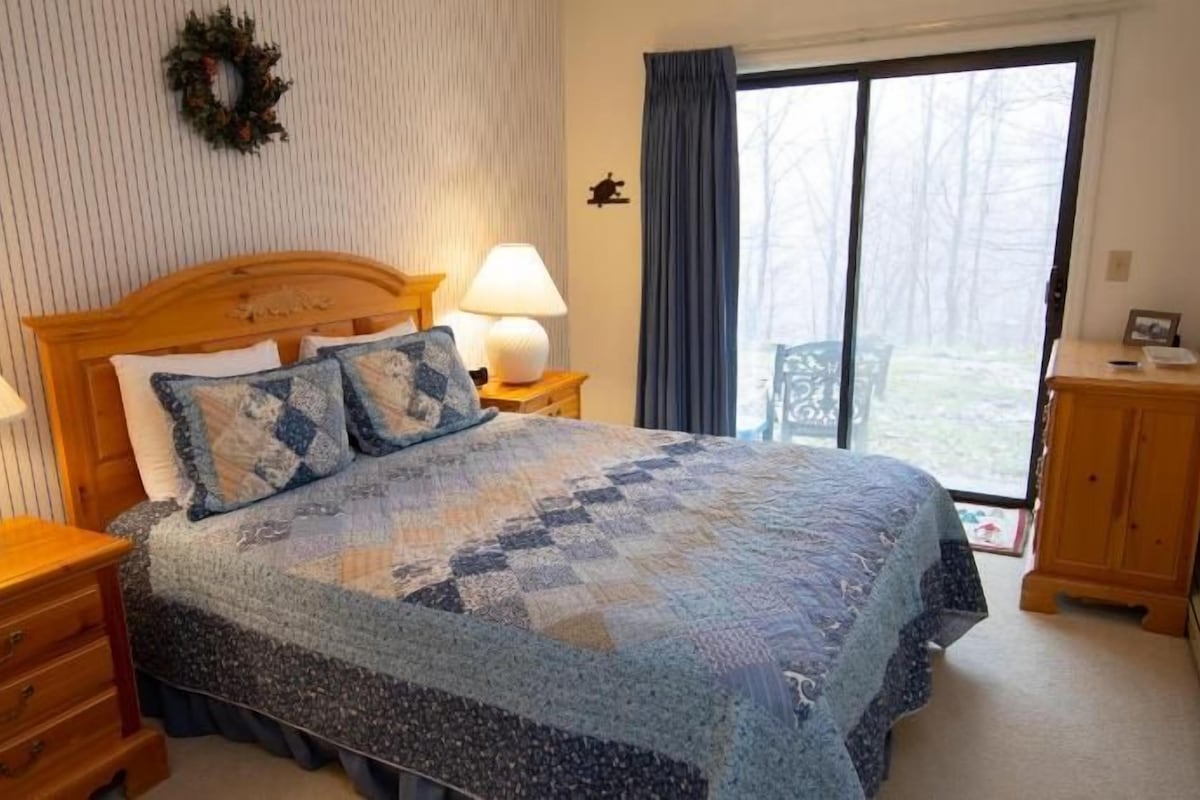 Une chambre à coucher comprenant un lit, deux tables de chevet, une commode, une coiffeuse, une fenêtre avec des rideaux et une couronne décorative accrochée au mur.