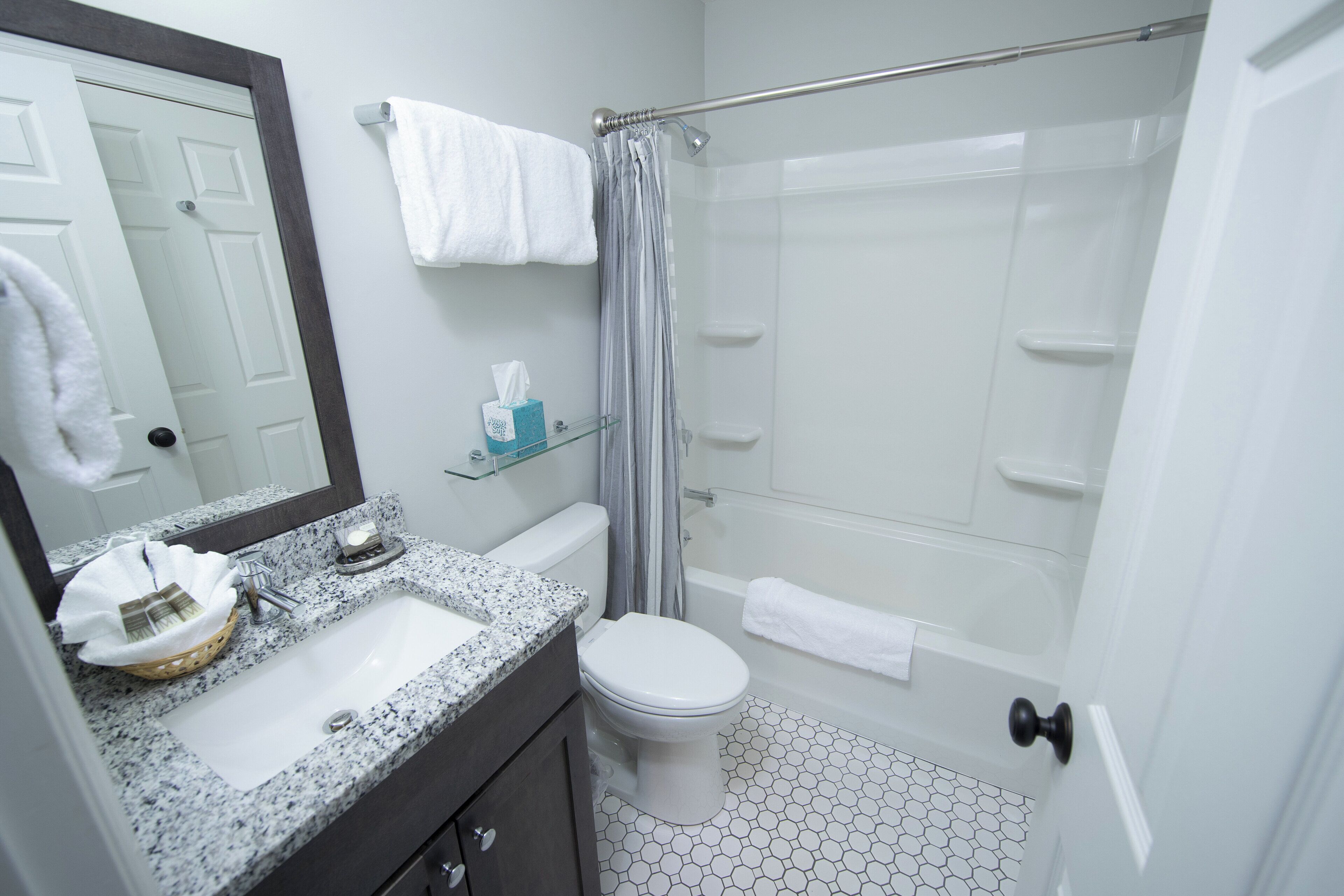 Un baño con lavamanos, inodoro y ducha blancos. Hay un perchero sobre el lavamanos y un estante de vidrio con artículos de aseo. El suelo tiene un diseño de baldosas hexagonales.