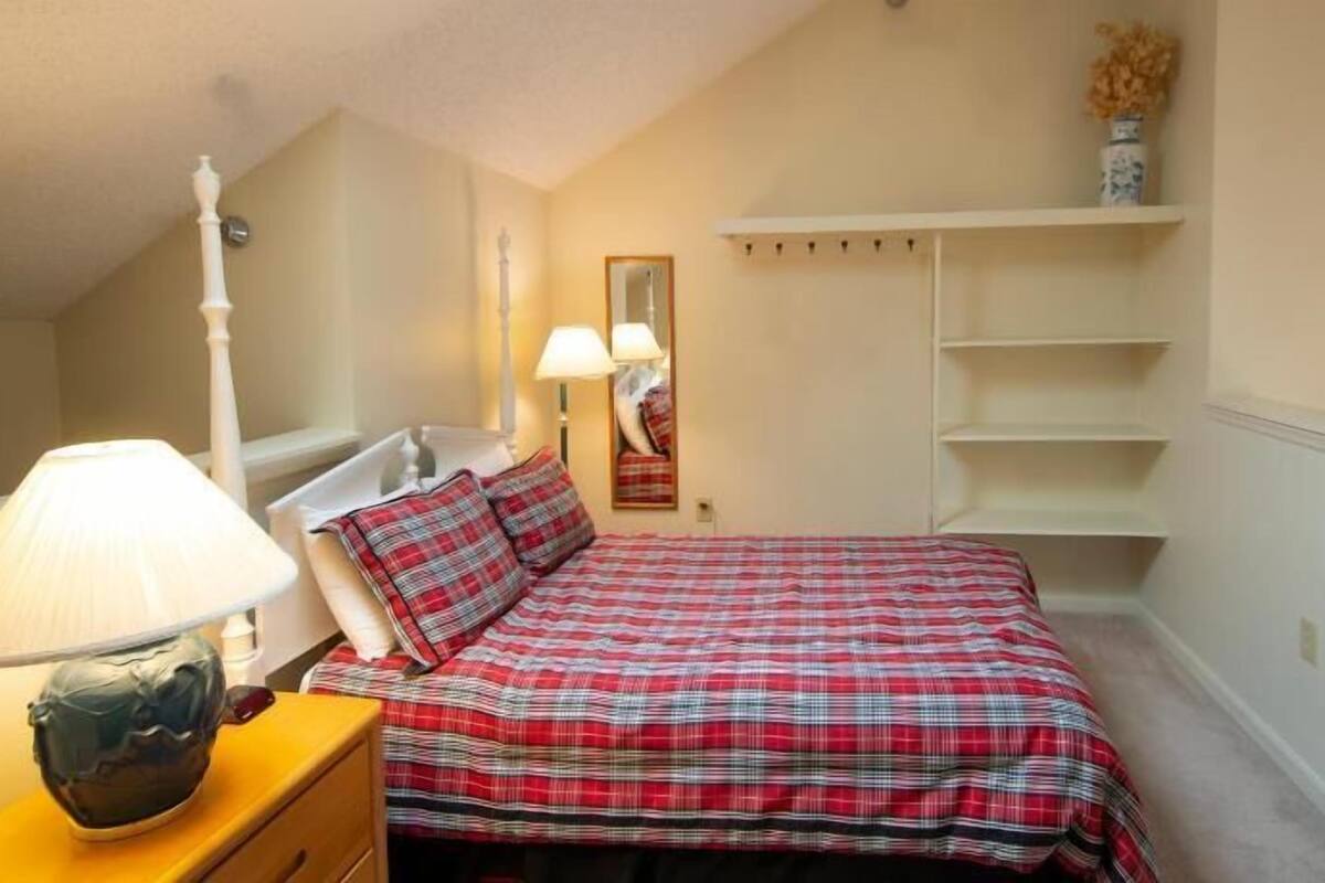 A bedroom with a bed, a nightstand, a lamp, a mirror, and a shelf with a vase.