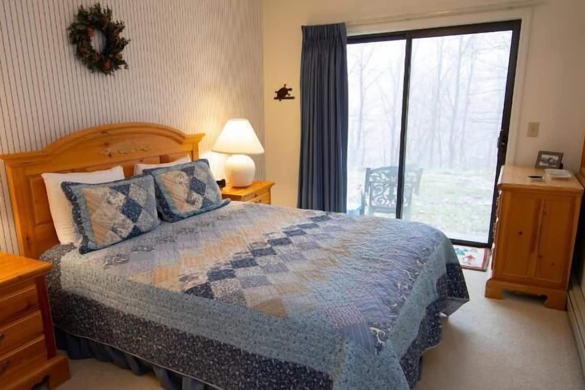 A bedroom with a bed, two bedside tables, a nightstand, a dresser, a window with curtains, and a wall-mounted wreath.