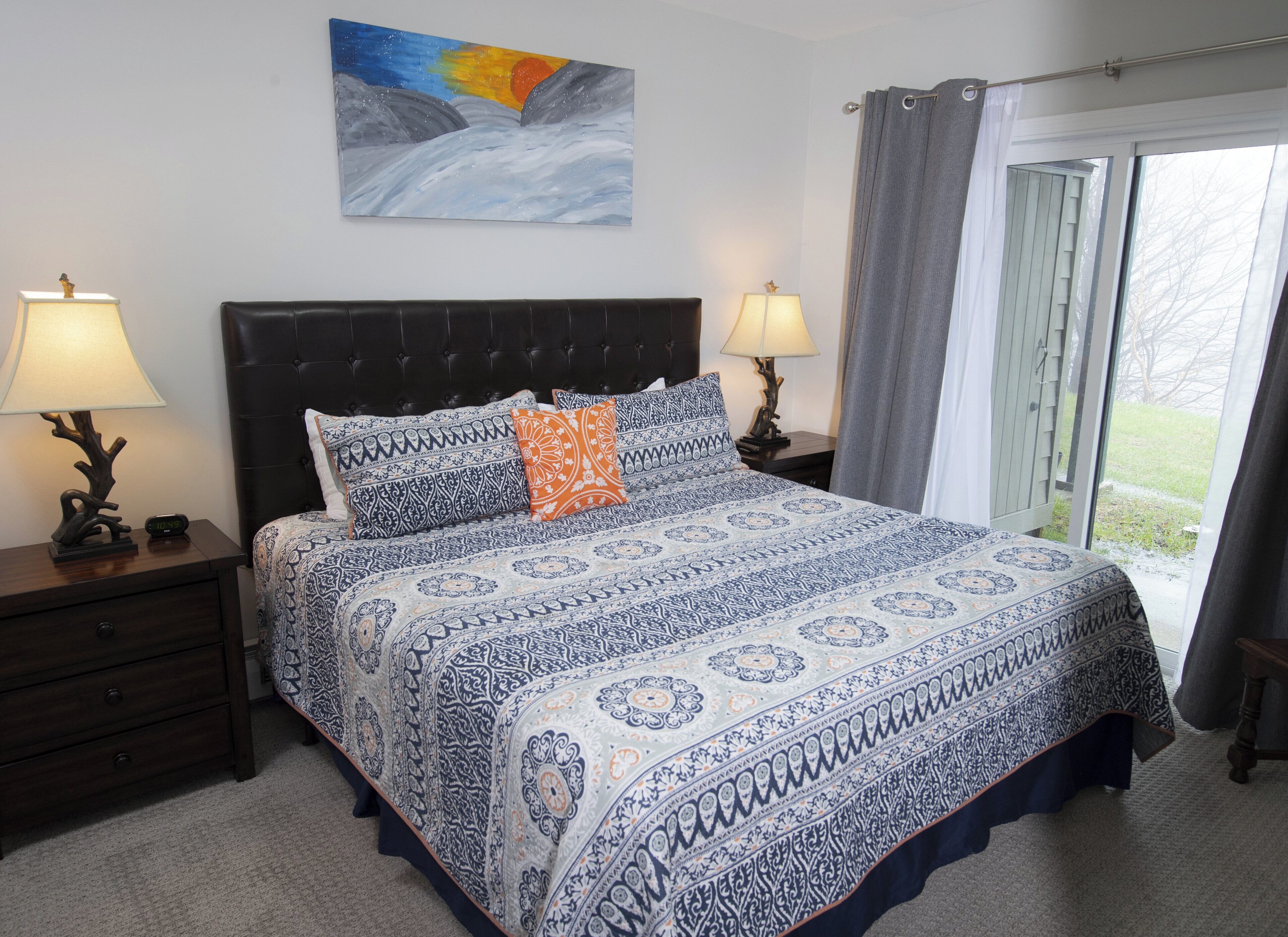 A bedroom with a large bed, two bedside tables with lamps, a nightstand with a lamp, a painting on the wall, and a view through a window with curtains.
