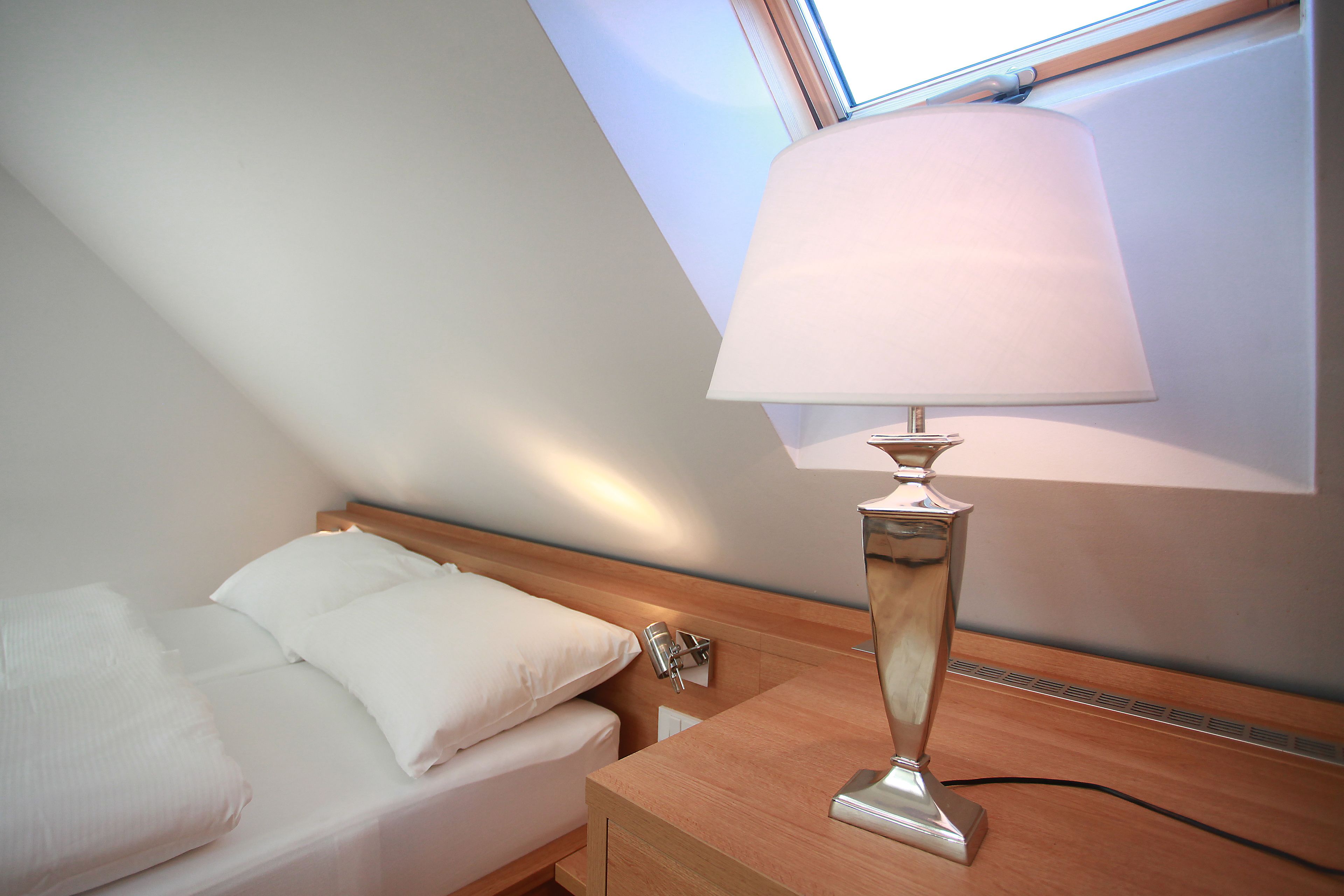 Una cama bien tendida con sábanas blancas en una habitación con techo inclinado y tragaluz.