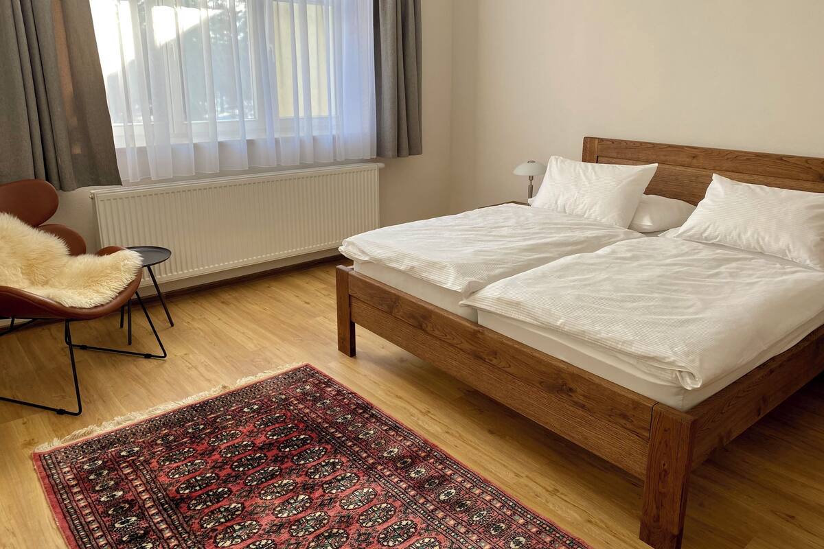 Un dormitorio con una cama de madera, un tapete rojo estampado, una silla y una ventana con cortinas.