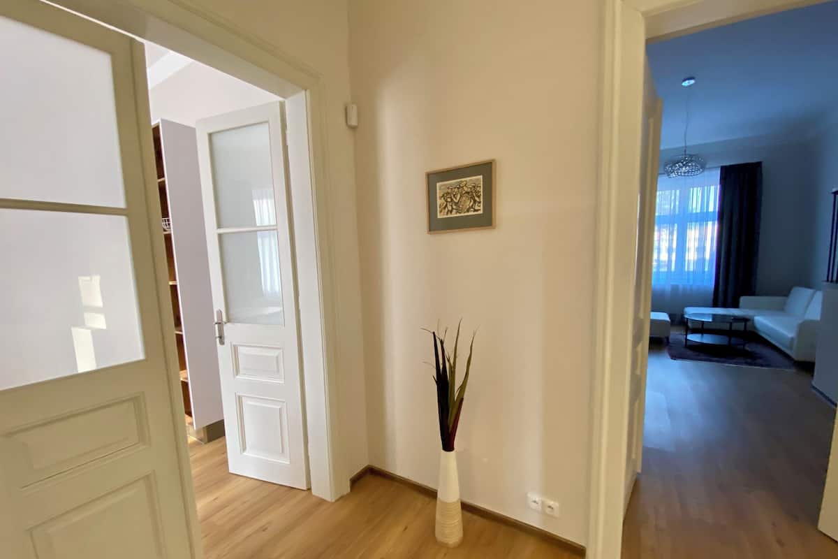 Una puerta abierta que da a un cuarto con piso de madera, un jarrón blanco con ramas y un cuadro en la pared.