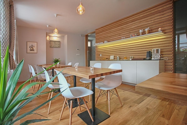A modern dining area with a wooden table and white chairs, a kitchen counter with appliances, and a hanging light fixture.