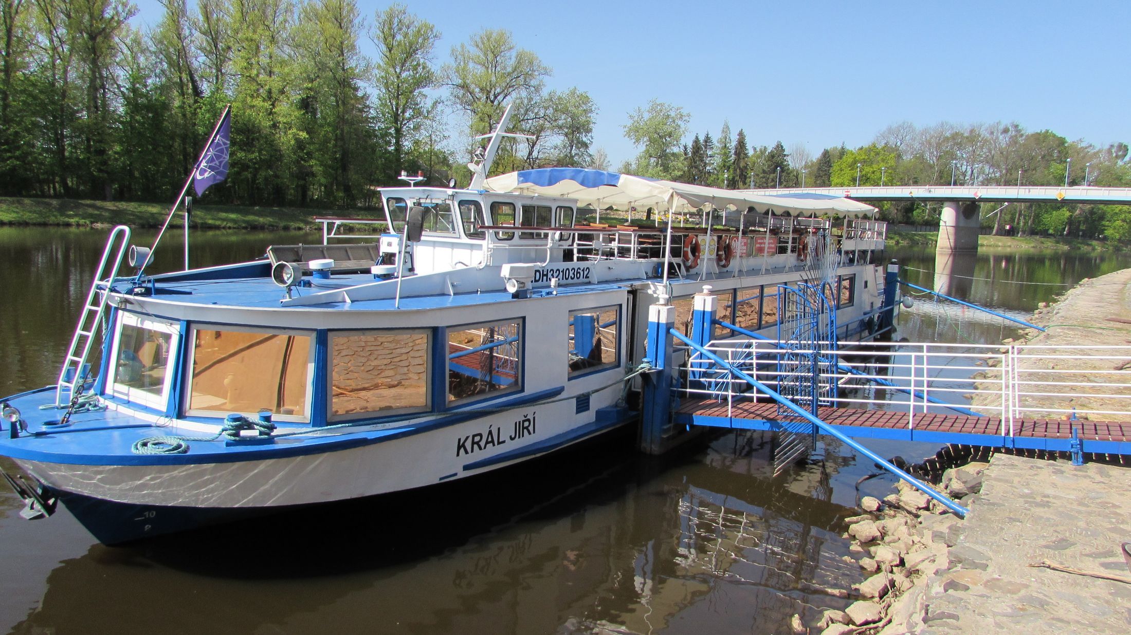 A riverboat docked at a pier with a covered deck area and a lifebuoy visible.