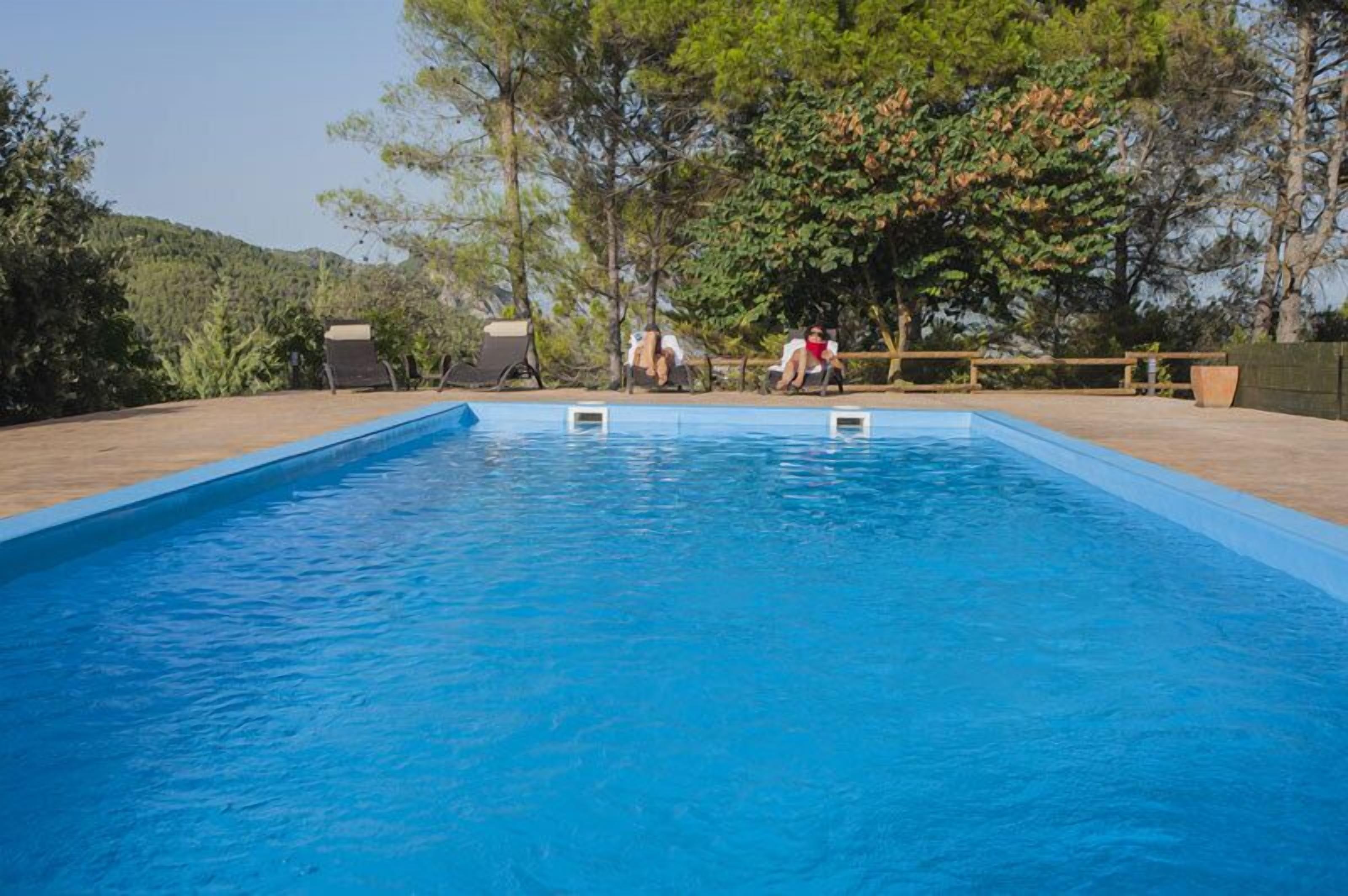 Una piscina rectangular con una terraza de madera alrededor. Se ven sillones de descanso y una valla de madera al fondo.