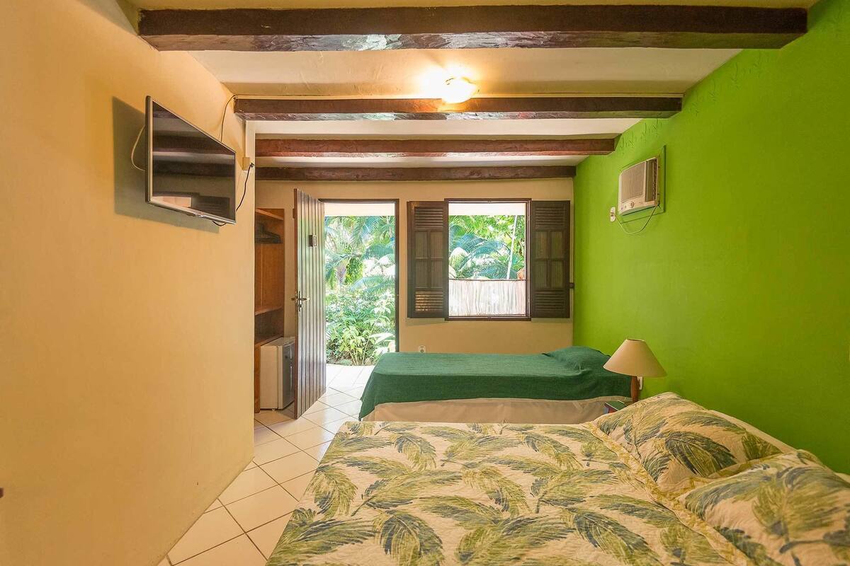 A bedroom with a green wall, a bed with a leaf-patterned bedspread, a TV mounted on the wall, and a window with shutters.