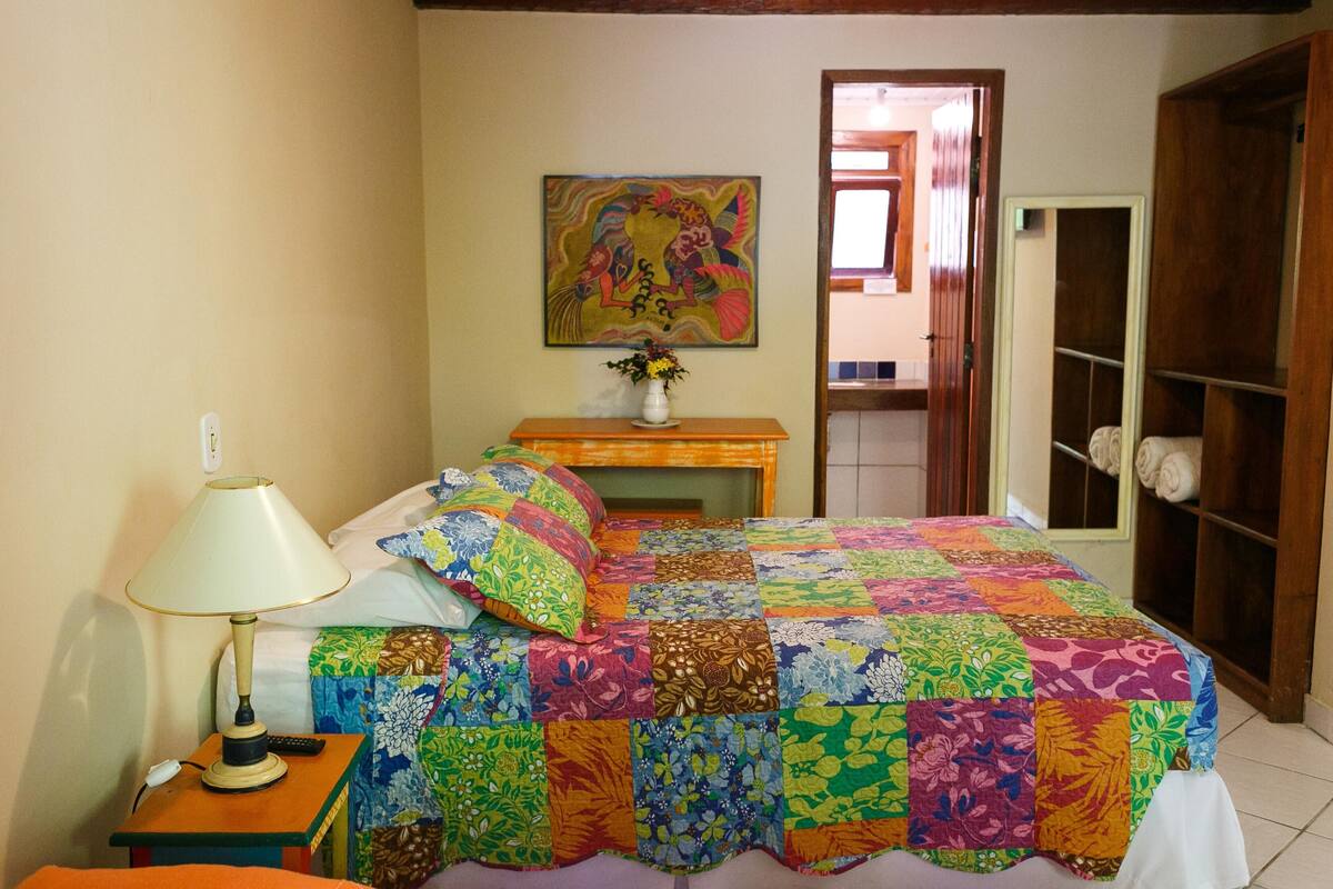 A bedroom with a colorful quilt, a bedside table with a lamp, a painting on the wall, and a wooden wardrobe.