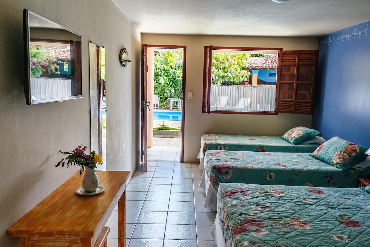 A room with two beds, a wooden table with a vase of flowers, and a view of a blue pool and greenery outside.