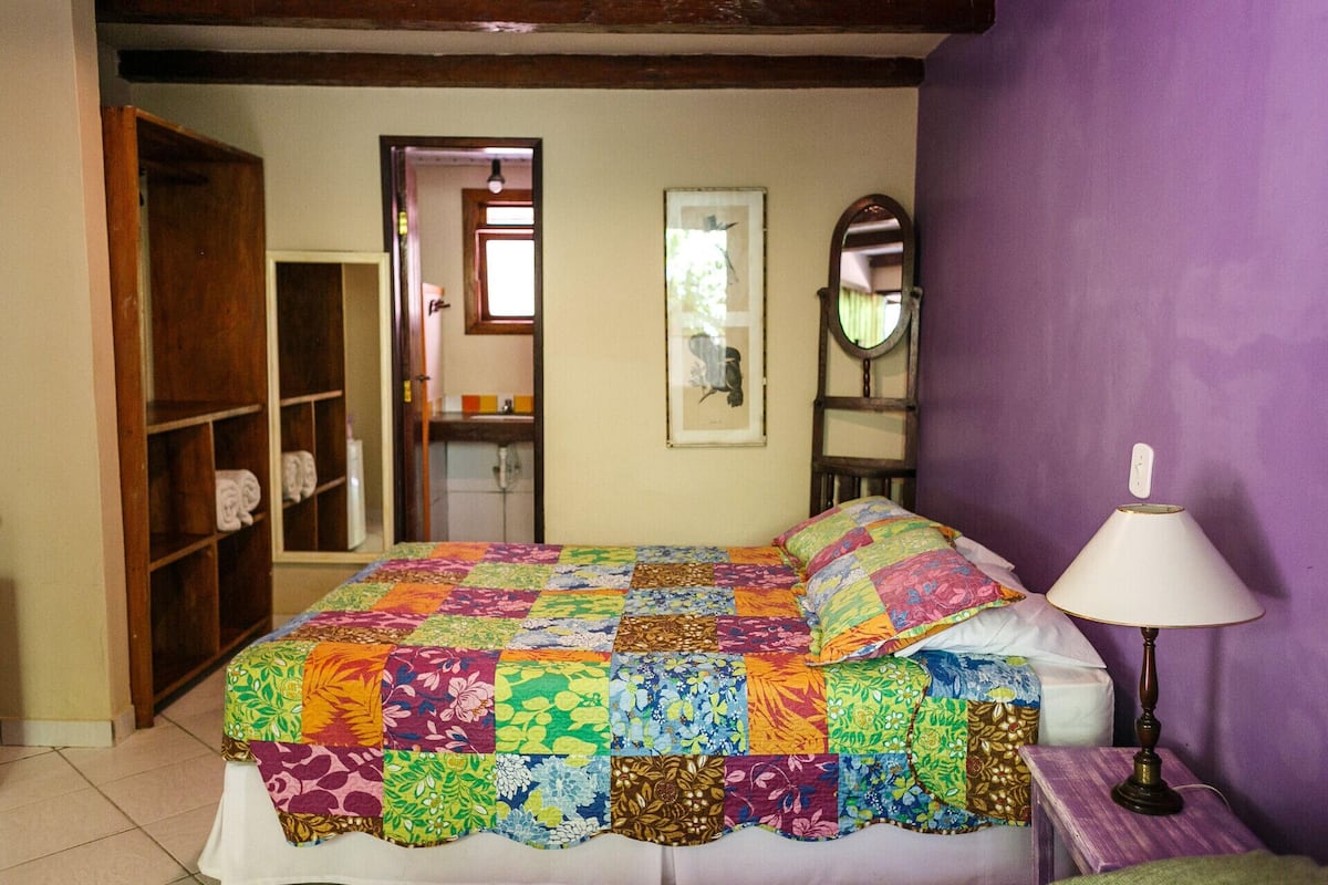 A bedroom with a colorful quilted bedspread, a wooden wardrobe, a mirror, and a bedside lamp.