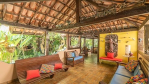 A covered outdoor seating area with wooden benches and colorful cushions, surrounded by lush greenery and a tiled floor. - Pousada Bemvirá (Porto Seguro)