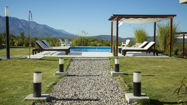 Een zwembad met ligbedden en een pergola, omringd door een grindpad en groen, met bergen op de achtergrond.