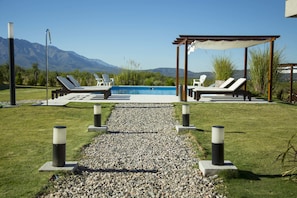 Zona de piscina con sillones de descanso y una pérgola, rodeada por un sendero de grava y vegetación, con montañas al fondo.