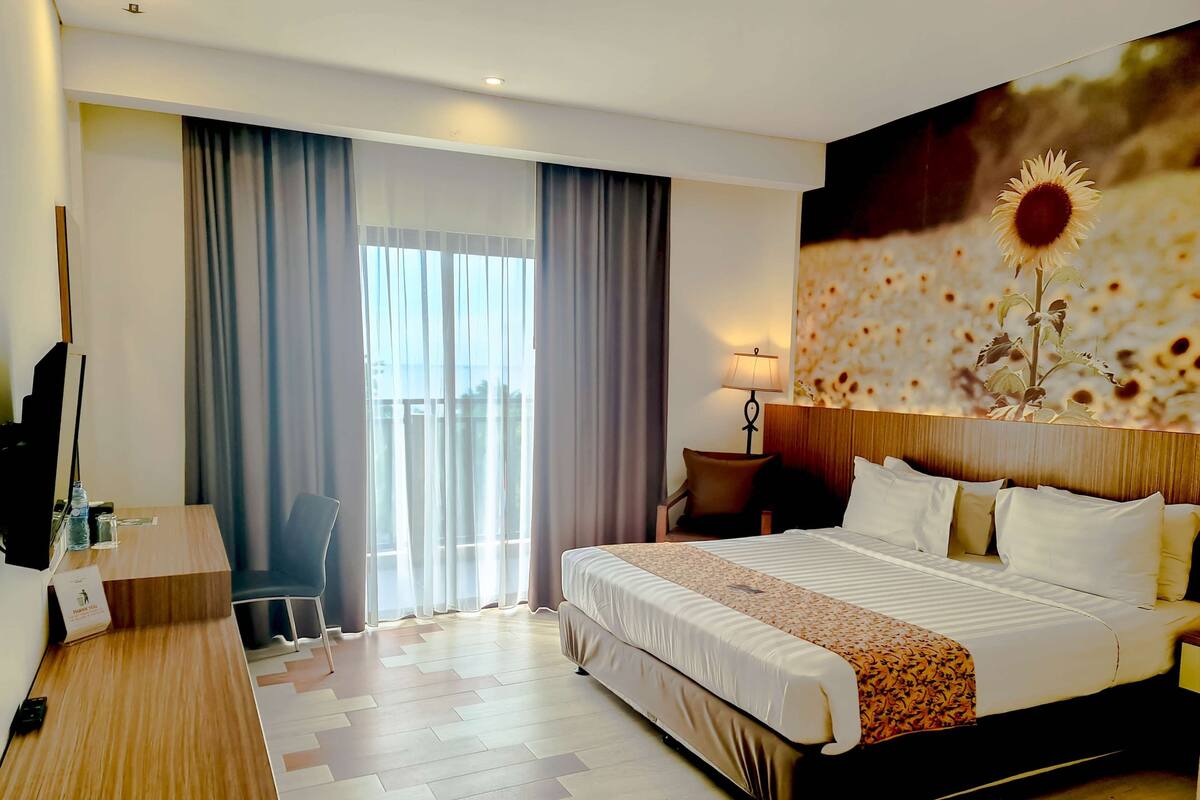 A hotel room with a large bed, a desk, a chair, a lamp, and a view of the outdoors through a window.