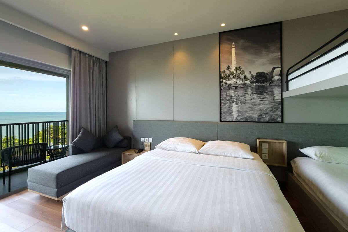 A modern hotel room with a large bed, a seating area with a view of the ocean, and a painting of a coastal scene.