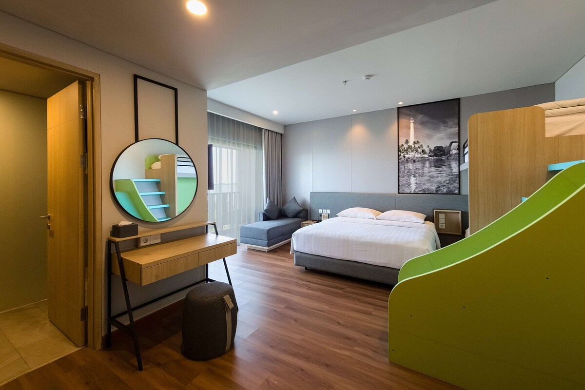 Een moderne hotelkamer met een bed, een bureau, een stoel, een spiegel en een groene glijbaan.