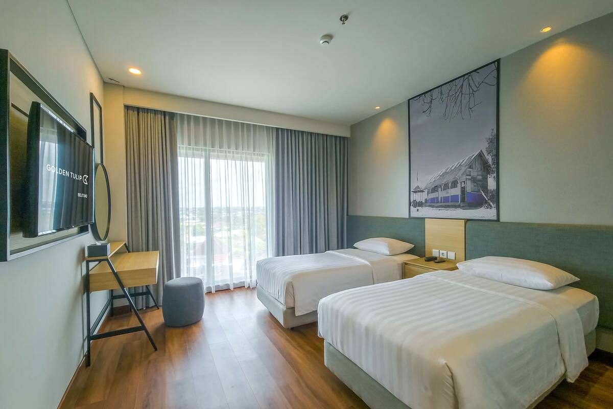 A hotel room with two beds, a TV, a desk, and a large window with curtains.